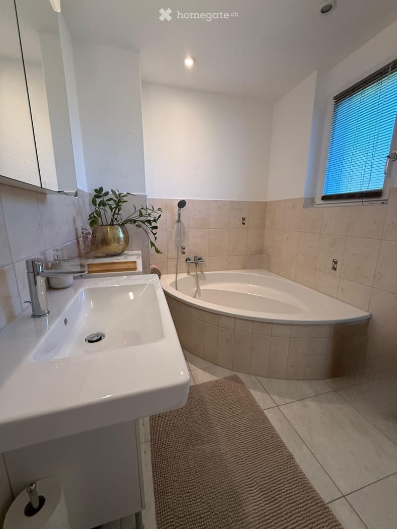 Modern bathroom with a corner bathtub, a large sink with a modern faucet, a toilet, a small plant on the windowsill, and a window with blinds.