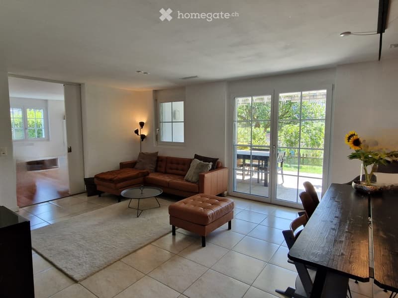 Modern living room with a brown leather sofa, ottoman, coffee table, dining table, and chairs. Large windows and sliding glass doors lead to a balcony.