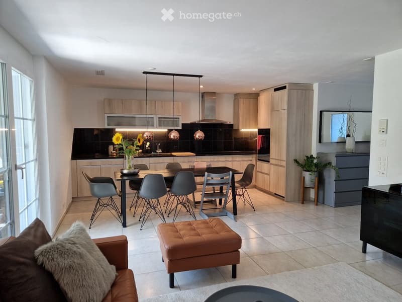 Modern open-plan living and dining area with a leather sofa, ottoman, dining table with chairs, kitchen with cabinets and island, pendant lights, and decorative elements.