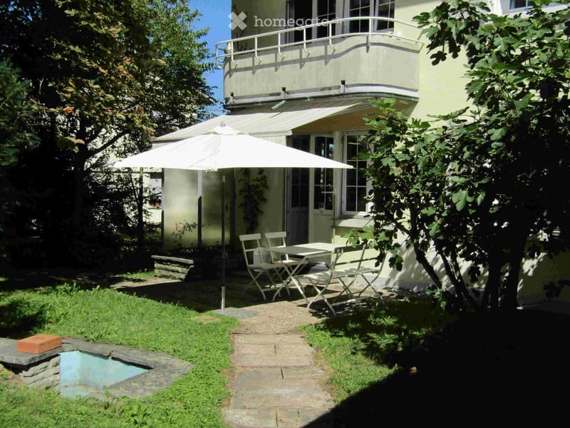 Garden area with a small swimming pool, a table with chairs, and an umbrella, surrounded by greenery and a house with a balcony.