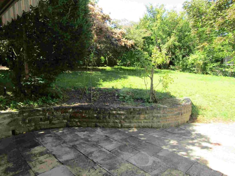 Garden area with a stone wall, paved patio, and lush greenery including trees and shrubs.