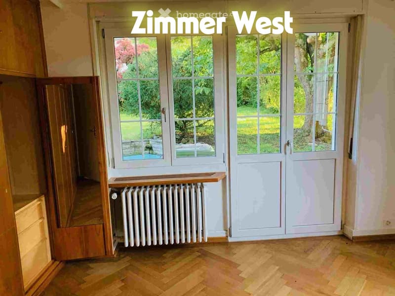 Empty room with wooden flooring, large windows, radiator, and a small wooden shelf near the window.