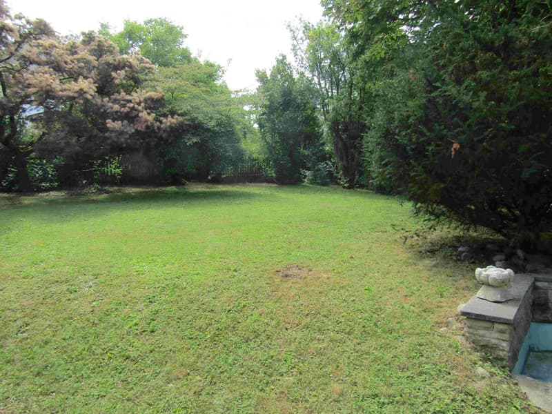 A well-maintained lawn surrounded by mature trees and shrubs, with a stone structure and small water feature visible on the right side.