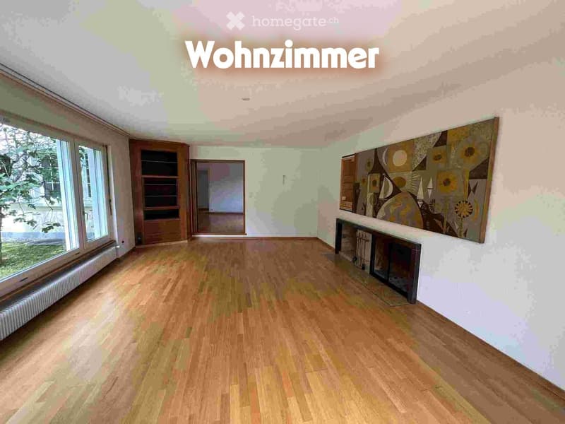 Empty living room with wooden flooring, large windows, and a fireplace. No furniture or furnishings are present.