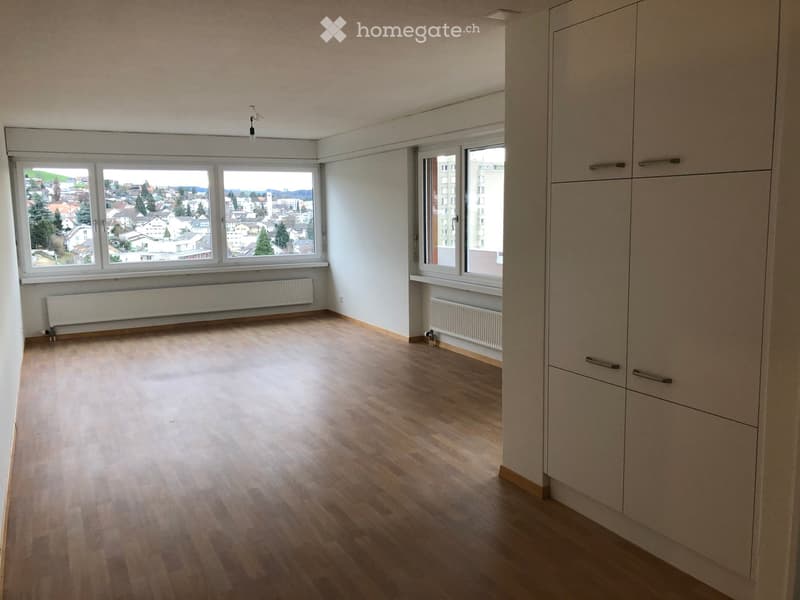 Empty room with wooden flooring, large windows offering a cityscape view, built-in closet, and radiators.