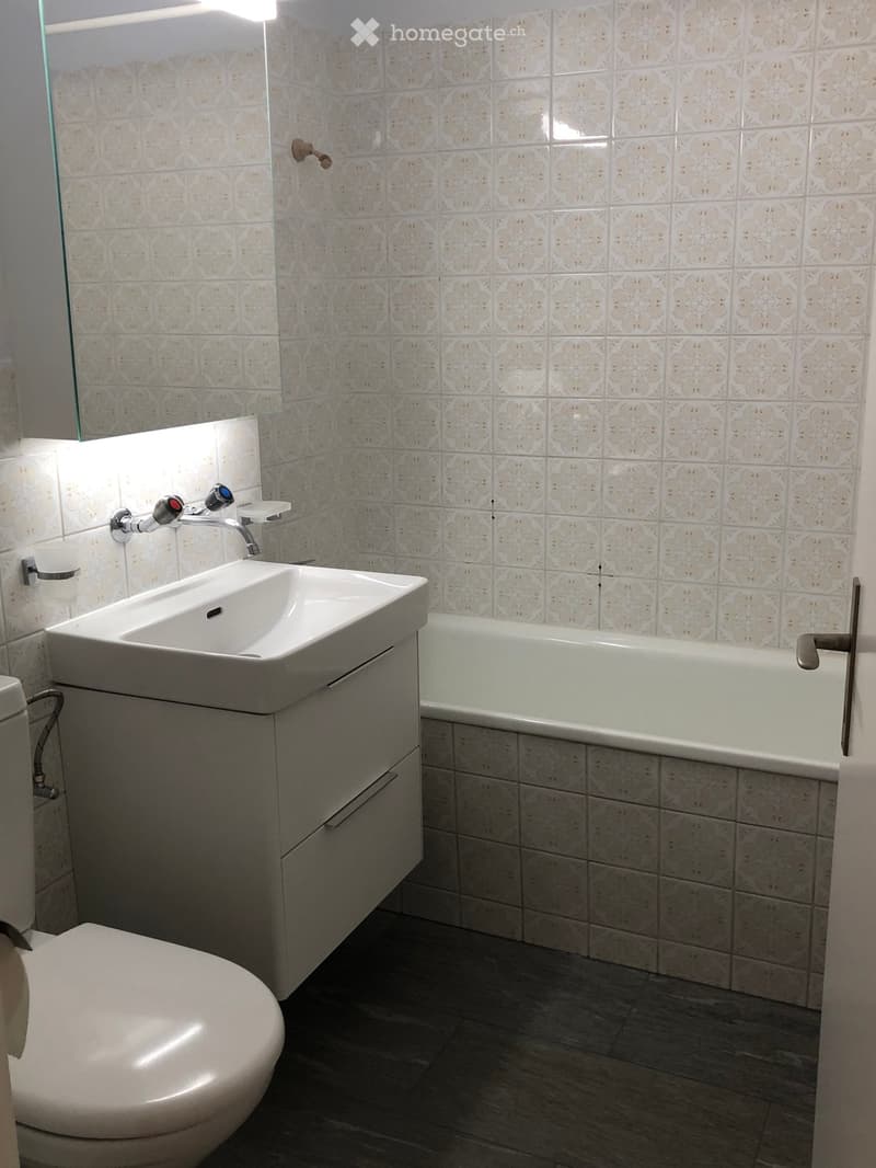 Modern bathroom with white tiles, a sink with a modern faucet, a bathtub, and a toilet. The walls are tiled with a decorative pattern, and the floor is dark grey.