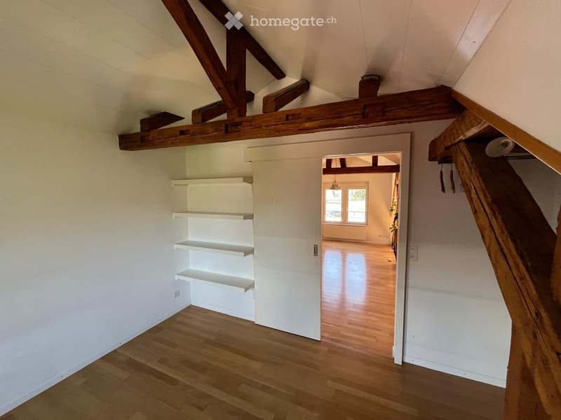 Empty attic space with wooden beams, white walls, wooden floor, and built-in white shelving unit. An open doorway leads to another room with wooden flooring and large windows.