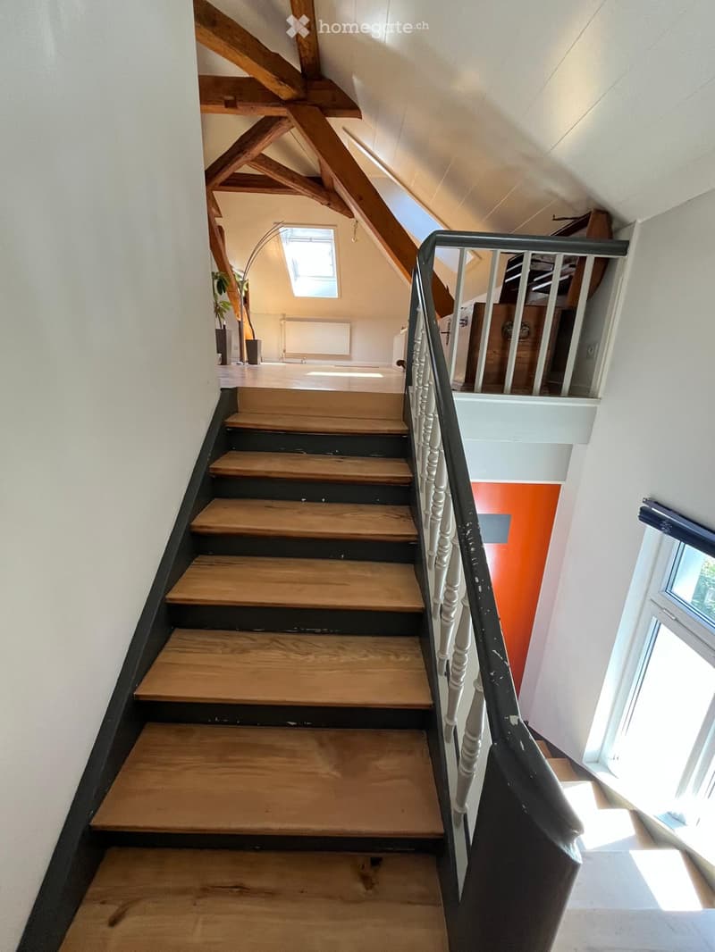 Wooden staircase with white balustrade, leading to an attic space with exposed wooden beams and a skylight window.