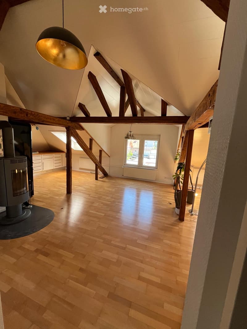 Bright, open attic space with wooden beams, wooden flooring, and large windows. Features a staircase leading to another level and a small cabinet area.