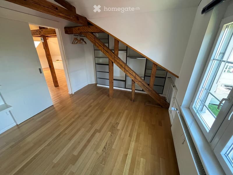 Empty hallway with wooden flooring, white walls, and a staircase leading upwards. A window on the right side allows natural light to enter.