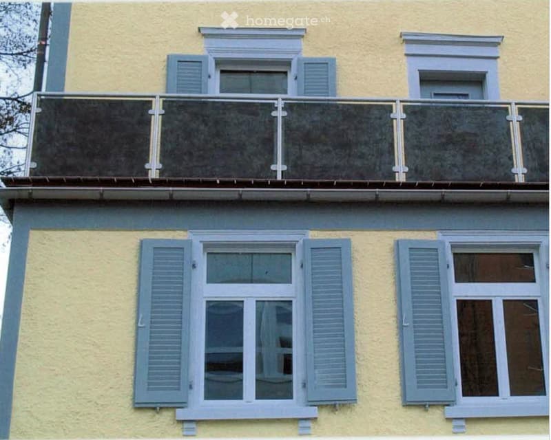 Yellow building facade with multiple windows, shutters, and a balcony with a glass railing.