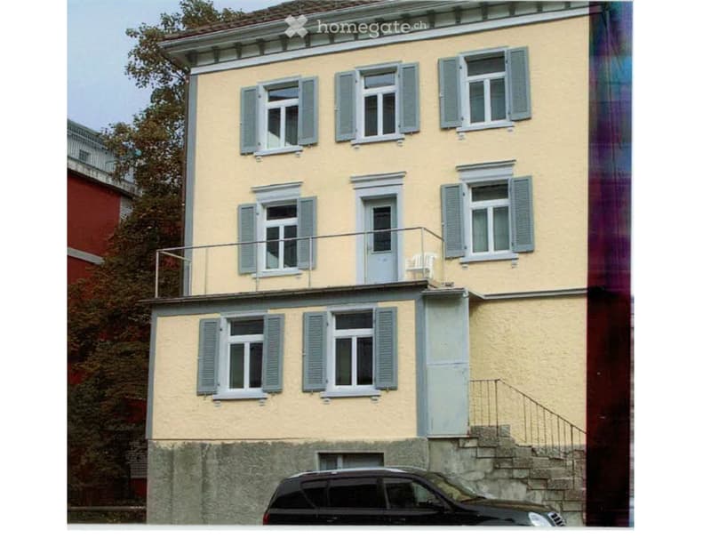 Three-story residential building with light yellow facade, multiple windows with shutters, and a balcony on the second floor.