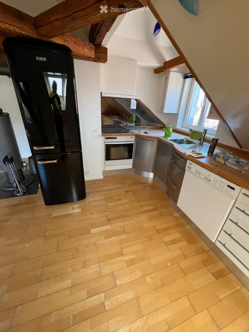Modern kitchen with wooden flooring, white cabinets, stainless steel appliances including a refrigerator, oven, and dishwasher, and a countertop with a sink and various items.