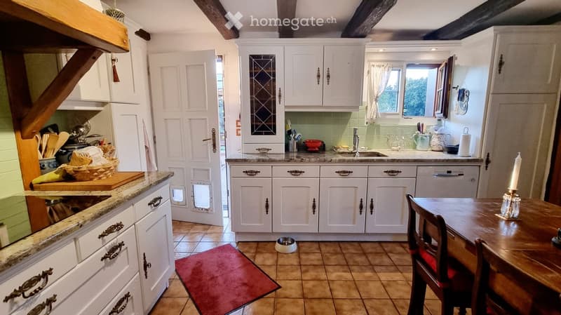 Bright kitchen with white cabinets, granite countertops, and a wooden dining table with chairs. Features include a built-in oven, sink, and various kitchen utensils.