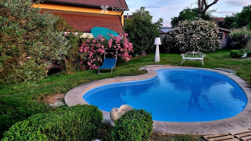 Well-maintained garden with a swimming pool, lounge chair, white bench, and decorative elements like a lamp and a shell sculpture.