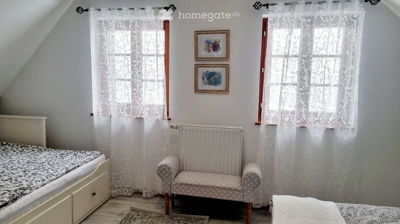 Cozy attic bedroom with wooden floors, white walls, large windows with lace curtains, a single bed with patterned bedding, a small armchair, a radiator, and framed artwork on the wall.