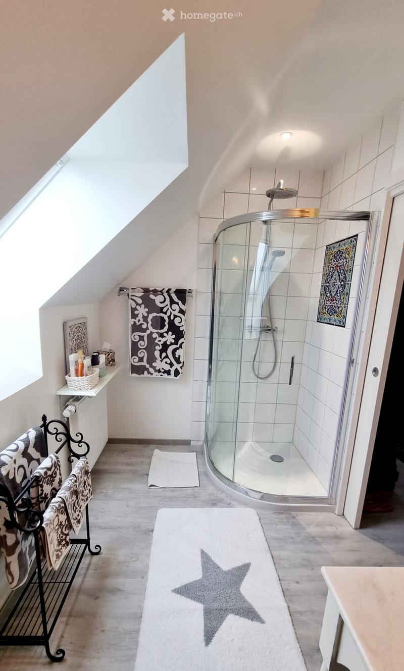 Modern bathroom with a glass-enclosed shower, white tiles, a towel rack with patterned towels, a white bath mat with a star design, and a shelf with toiletries.