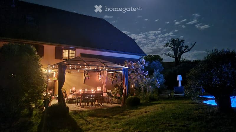 Well-lit outdoor patio area with a dining table, chairs, umbrella, and a swimming pool in the background.