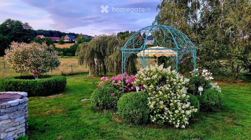 Well-maintained garden with a decorative gazebo, lush greenery, flowering bushes, and a stone pathway.