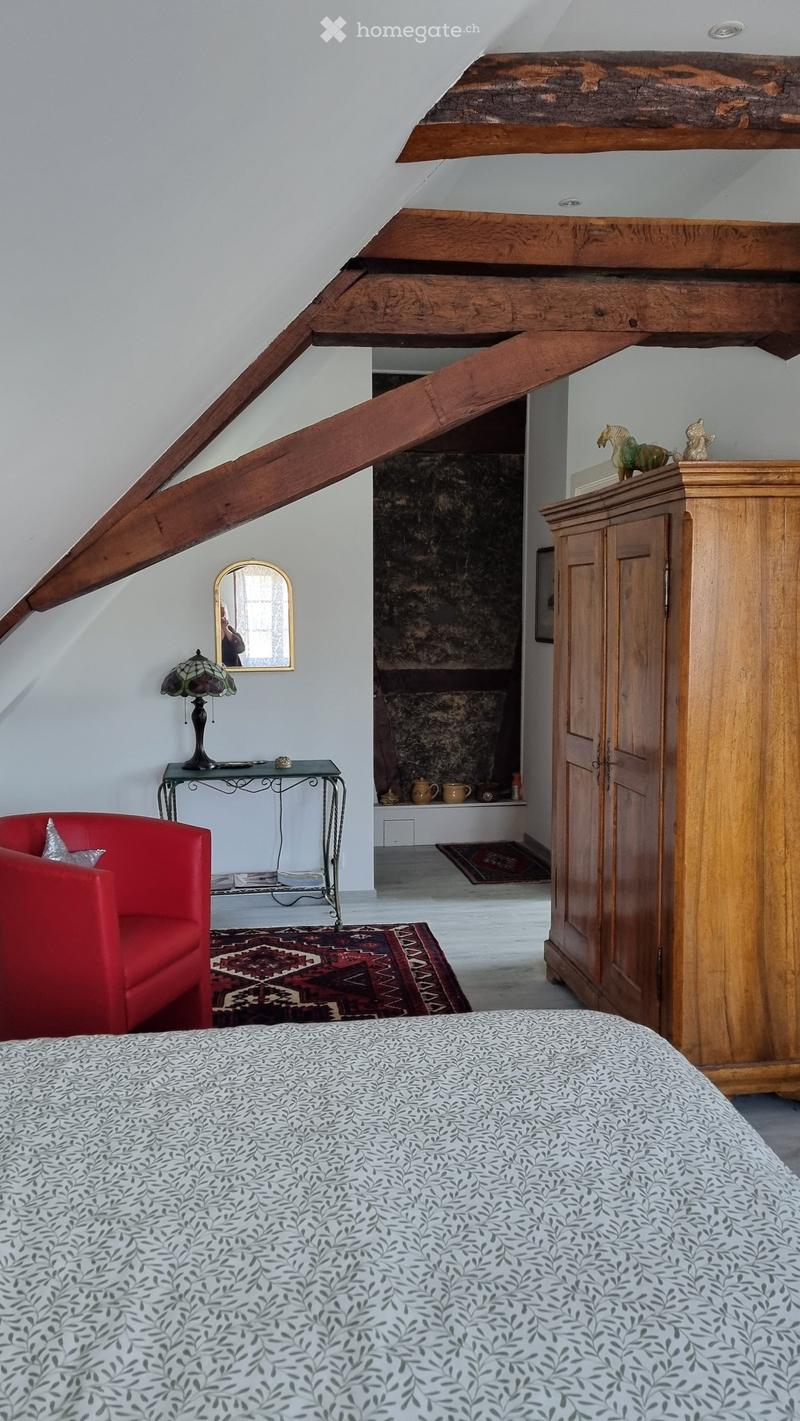 Cozy attic bedroom with exposed wooden beams, a patterned bedspread, a red armchair, a small table with a stained-glass lamp, and a wooden cabinet with decorative items on top.