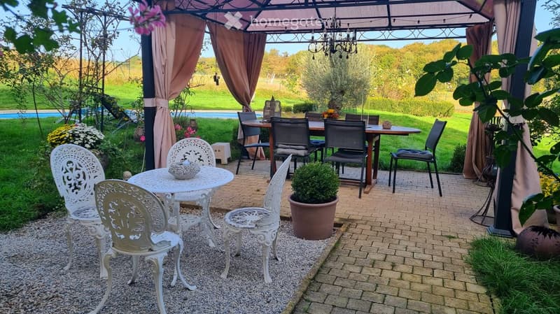 Outdoor patio with a covered seating area, featuring a white metal table and chairs, a larger wooden dining table with chairs, and a potted plant. The area is surrounded by greenery and a swimming pool is visible in the background.