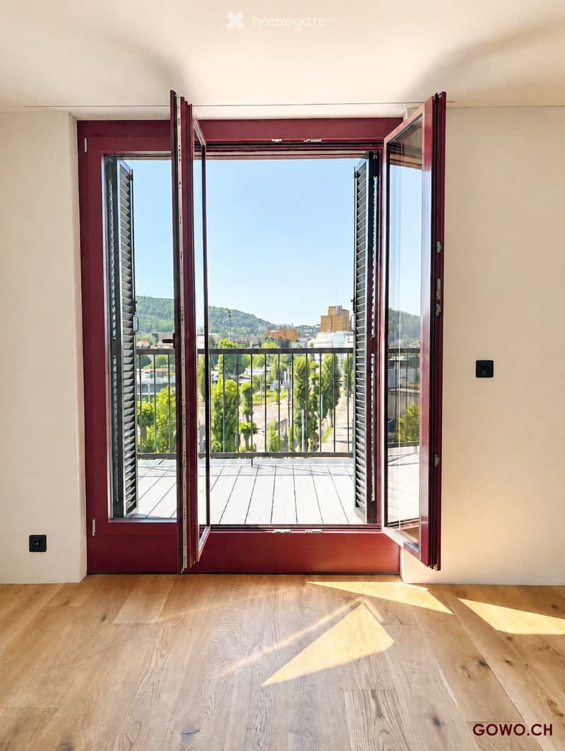 Empty room with wooden flooring, large open windows leading to a balcony with a city view.