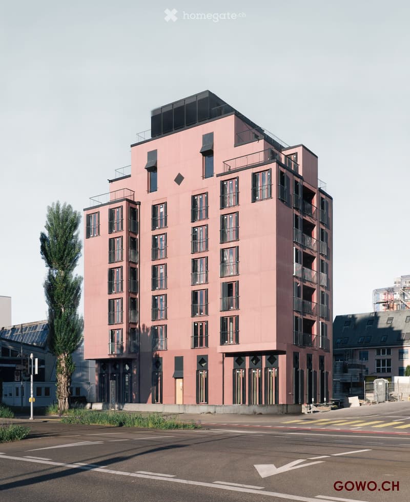 Modern multi-story building with pink facade, large windows, and balconies. Located near a road with visible parking area and greenery.