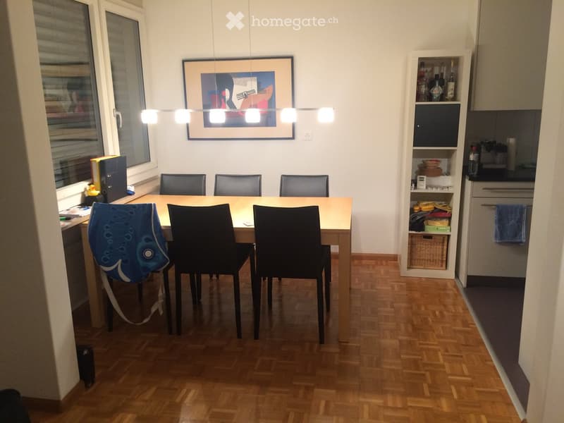 Dining area with a wooden table, six chairs, a shelf with bottles and items, and a framed picture on the wall.