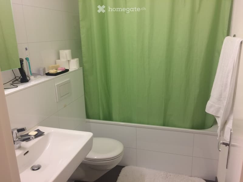 Modern bathroom with white tiles, a bathtub with green shower curtain, a sink with a faucet, a toilet, and a towel on the floor.