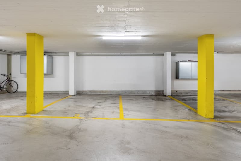 Empty underground parking area with concrete floor, yellow pillars, and marked parking spaces. A bicycle is positioned against the wall.