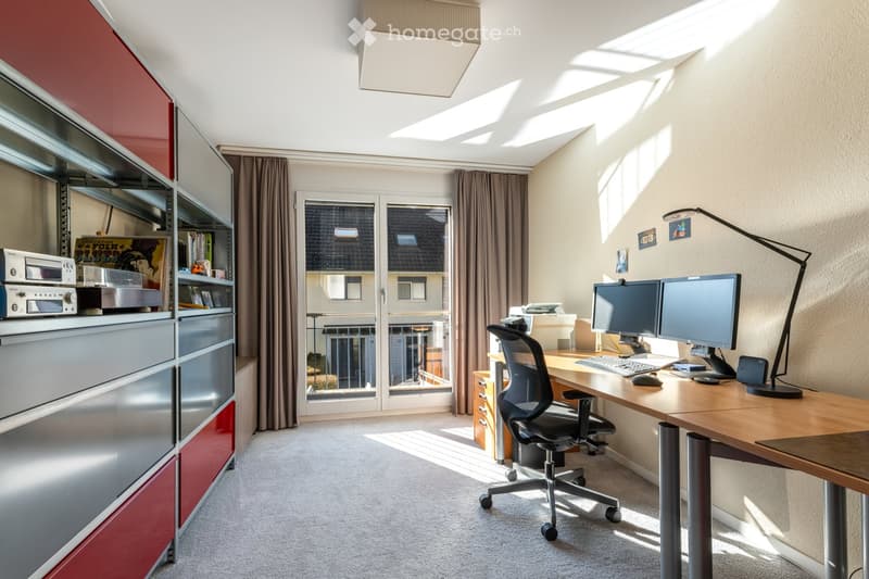 Modern, well-lit room with a large wooden desk, two computer monitors, ergonomic chair, floor lamp, and built-in red and grey cabinetry. Large window with curtains offers a view of the outside.