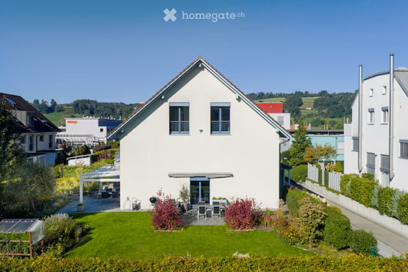 Single-family house with a modern design, white exterior, multiple windows, and a well-maintained garden with outdoor seating area.