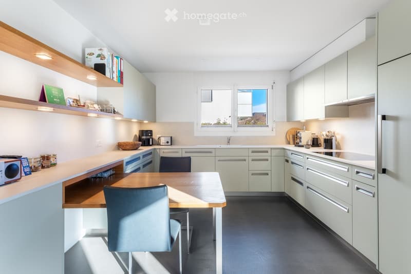 Modern kitchen with light grey cabinets, a wooden island, and a dining area. Features include a microwave, coffee maker, and various kitchen items on the counter and shelves.