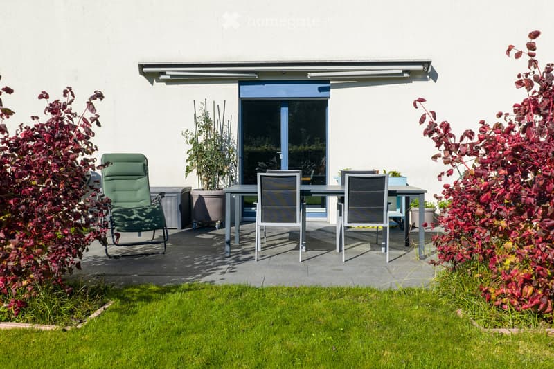 Outdoor patio area with a table, chairs, and surrounding greenery, connected to a house via a sliding glass door.