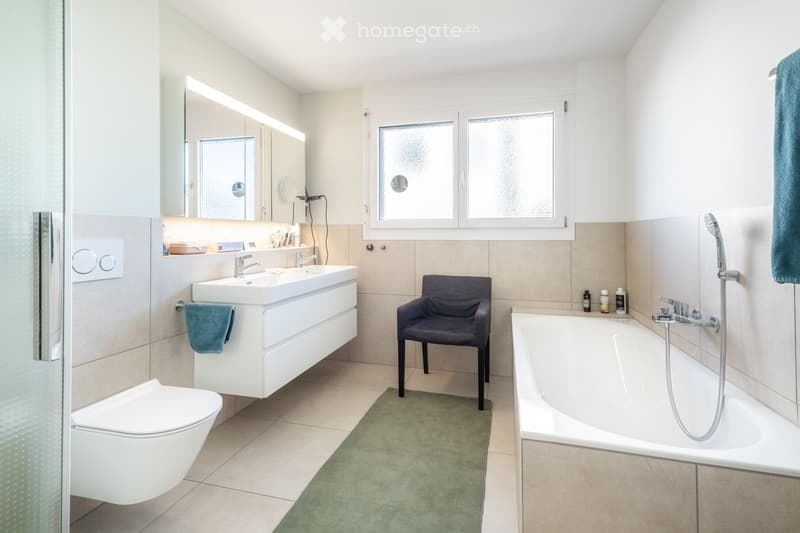 Modern bathroom with a white sink, mirror, toilet, bathtub, and a small dark grey chair. Beige tiles and a green bath mat are visible.