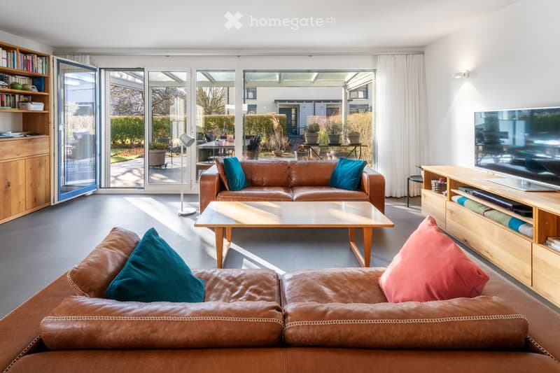 Bright living room with large glass doors leading to a patio, leather sofas, a coffee table, a TV stand, and a bookshelf.