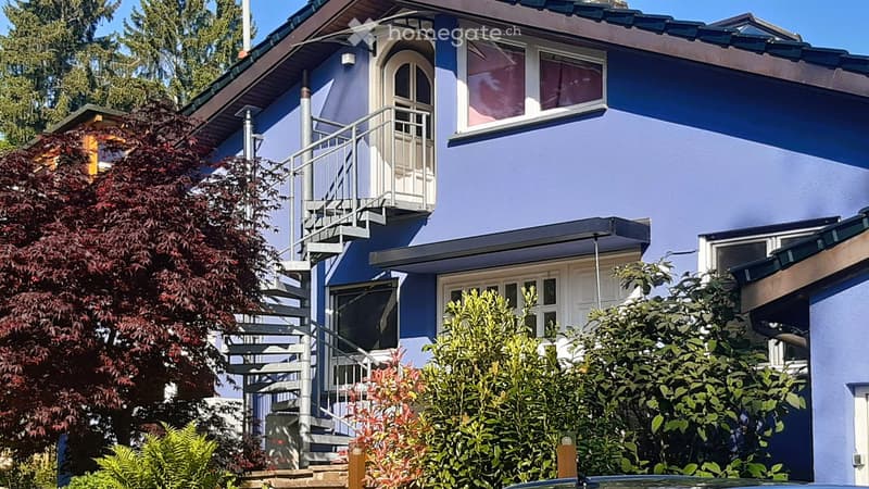Two-story house with blue exterior, white trim, and a balcony accessible via an external staircase. The house features multiple windows and is surrounded by greenery, including a red-leaved tree and shrubs.