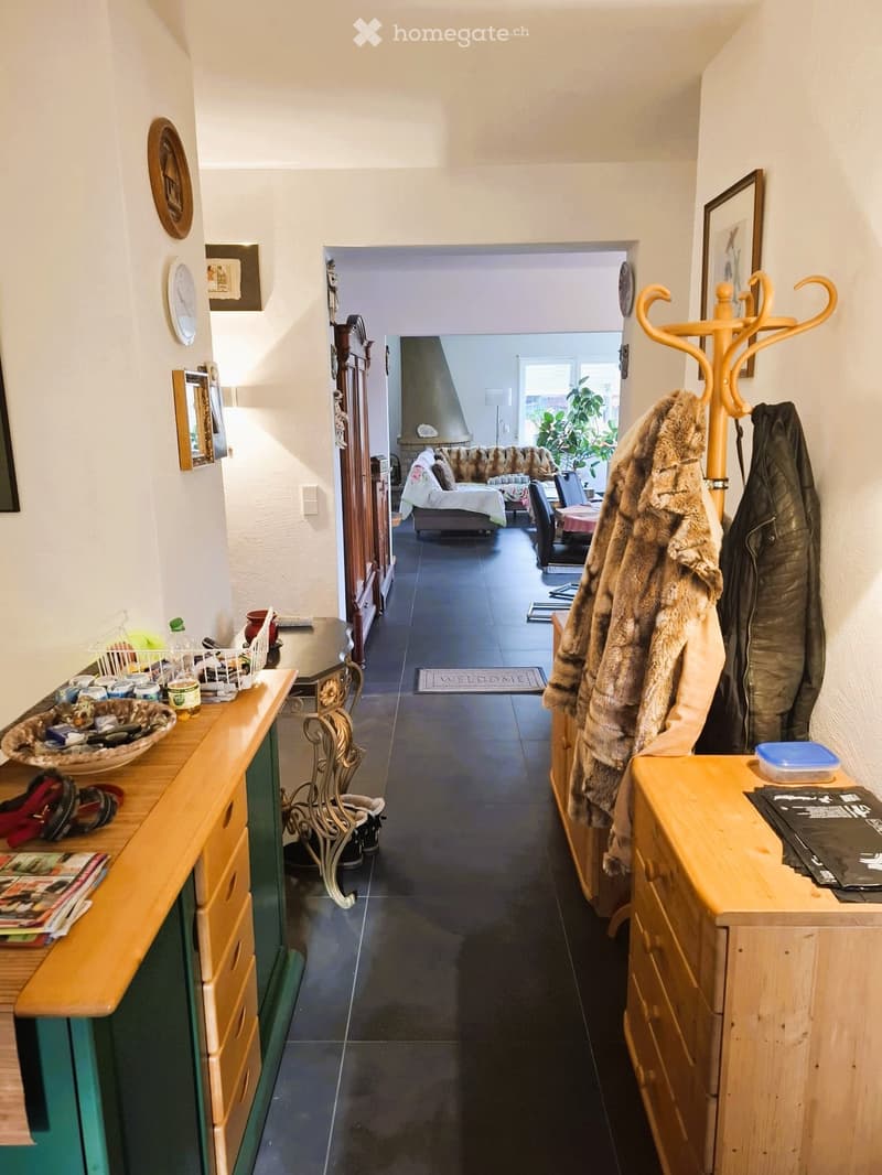 Entrance hall with wooden cabinets, coat rack, and a rug. The hallway leads to a living room with a couch, table, and a window.