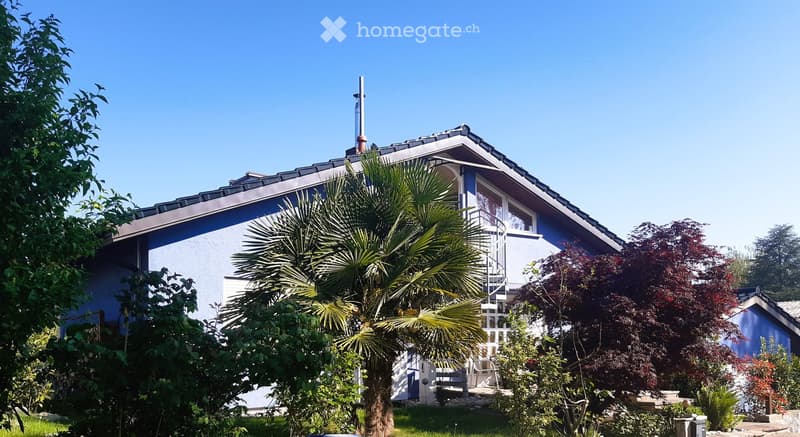 A two-story house with a blue exterior and white trim, featuring a spiral staircase leading to a balcony, surrounded by lush greenery and palm trees.