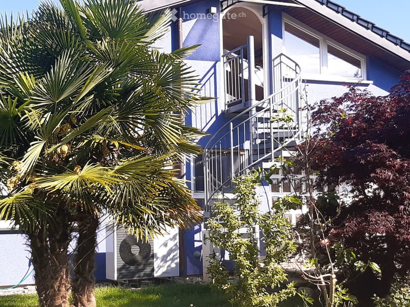 Two-story house with blue and white exterior, featuring a staircase leading to a balcony, surrounded by palm trees and shrubs.