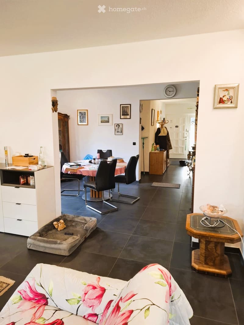 Living room with dining area, dining table with chairs, white walls, wooden cabinet, framed pictures, clock, wooden side table, rug, couch with floral cover