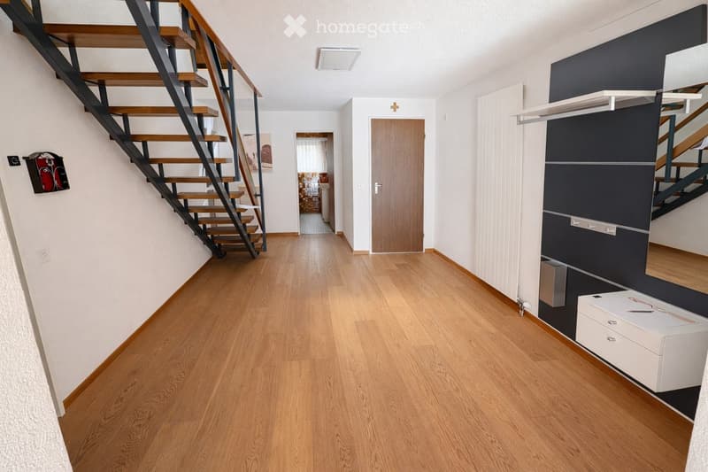 Modern hallway with wooden flooring, white walls, and a staircase leading upwards. Features include a built-in storage unit, a small white cabinet, and a door leading to another room.