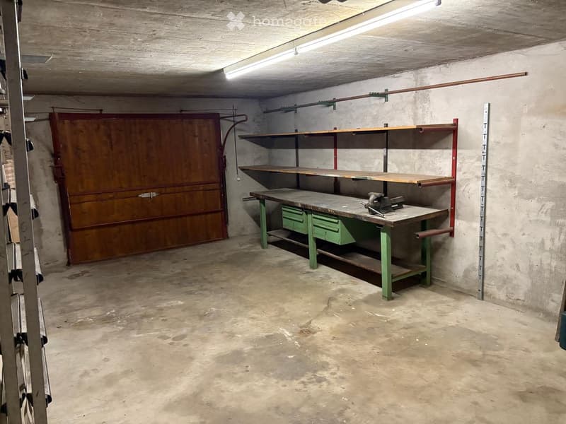 Empty workshop or garage with concrete floors, wooden workbench, shelves, and a large wooden door.