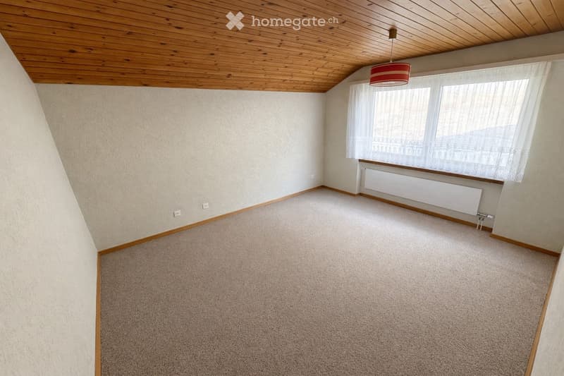 Empty room with beige carpet, light-colored walls, wooden trim, and a window with white curtains. A radiator is visible below the window, and a simple ceiling light fixture hangs from the wooden ceiling.