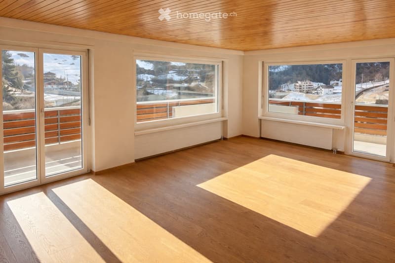 Empty room with wooden flooring, large windows, and sliding glass doors leading to a balcony. The room has a light-colored wooden ceiling and white walls.
