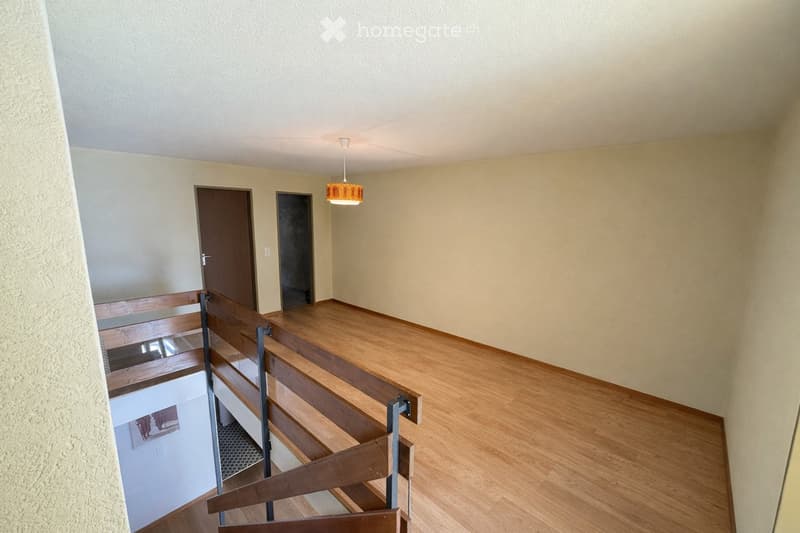 Empty hallway with wooden flooring, beige walls, and a hanging light fixture. A staircase leads downwards with wooden railings and a glass panel.