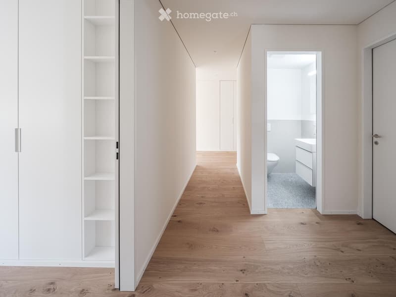 Empty hallway with wooden floor, white walls, and built-in shelves on the left side, leading to a bathroom with a toilet and sink.