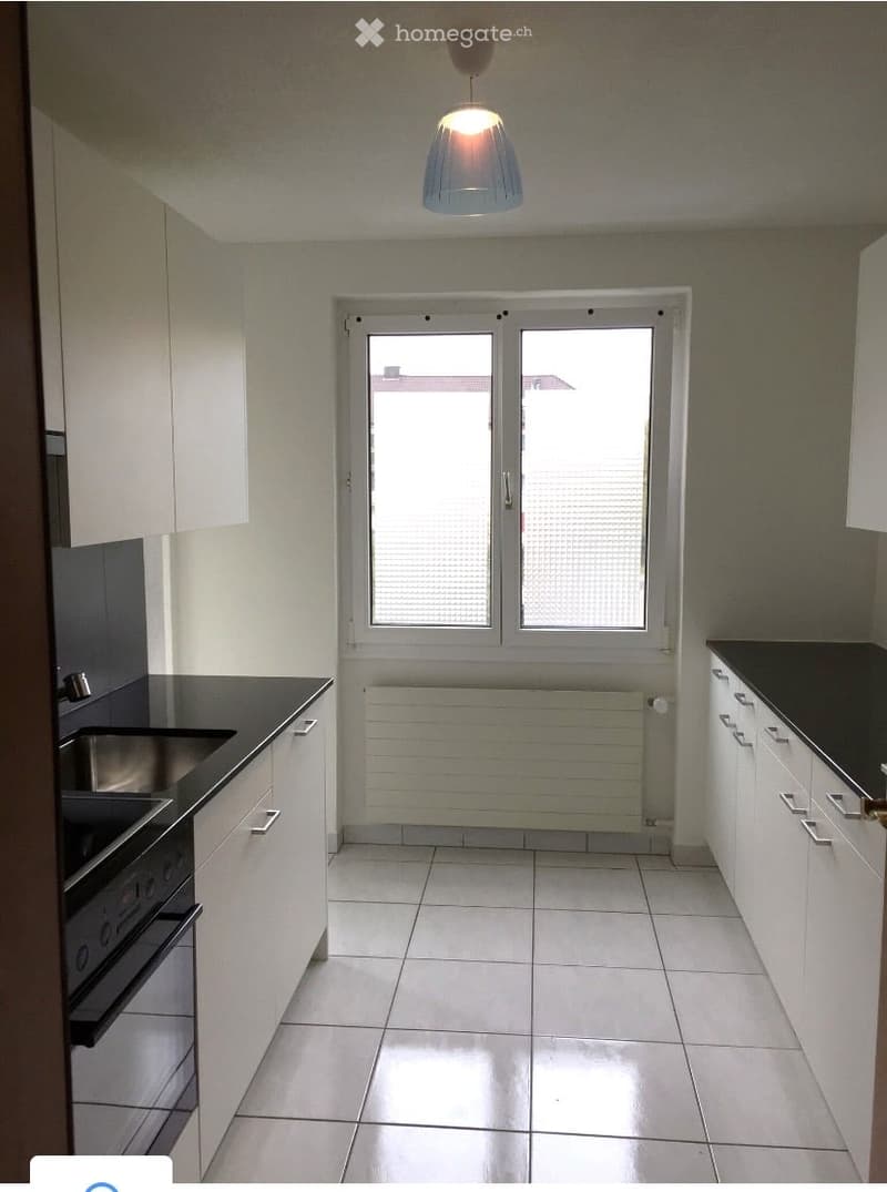 Modern kitchen with white cabinets, black countertops, and a stainless steel oven. The space features tiled flooring and a large window with blinds.