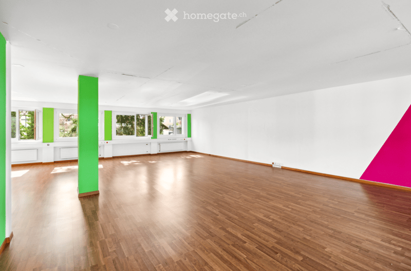 large open space with wood flooring, green pillars, white walls, windows with view of trees, bright natural light