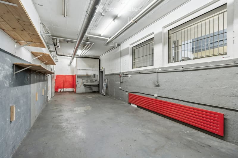 Empty hallway, mounted wooden shelves, metal piping on the ceiling, grey floor, red metal panel heater, glass barred windows